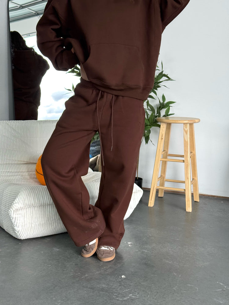 Person wearing a brown tracksuit in a room with a stool and plant.