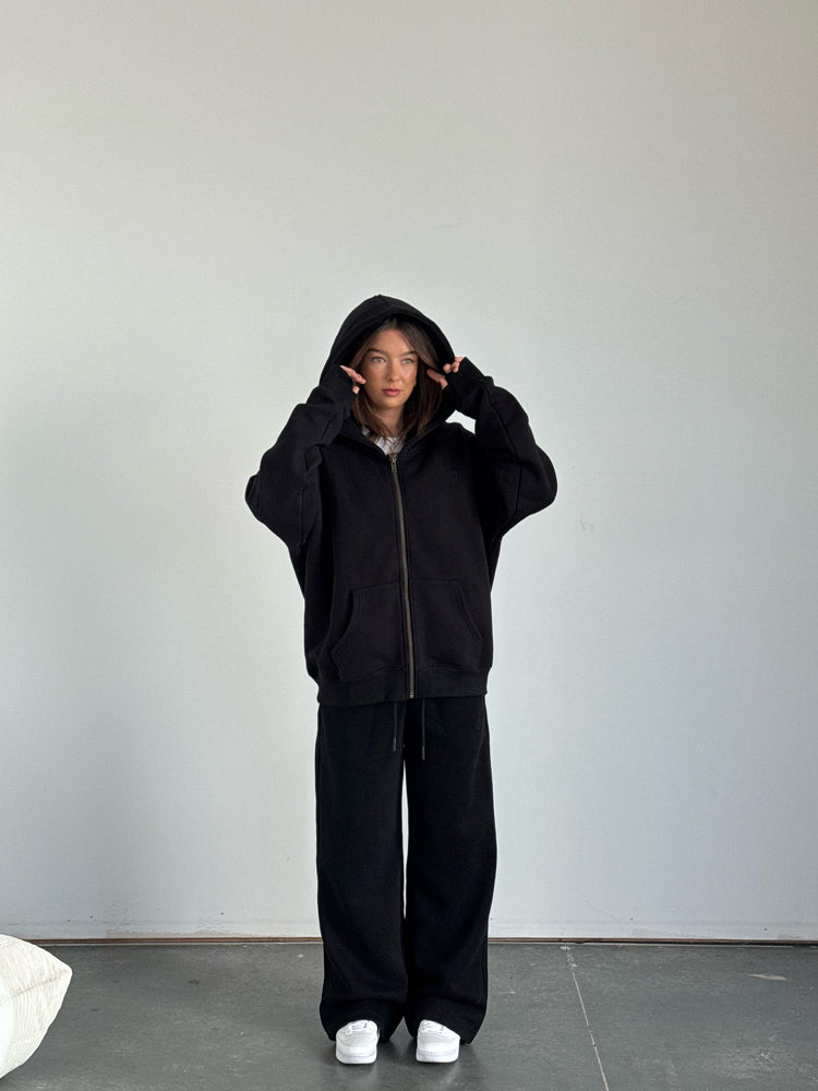 Person wearing a black hoodie and pants in a minimalistic setting