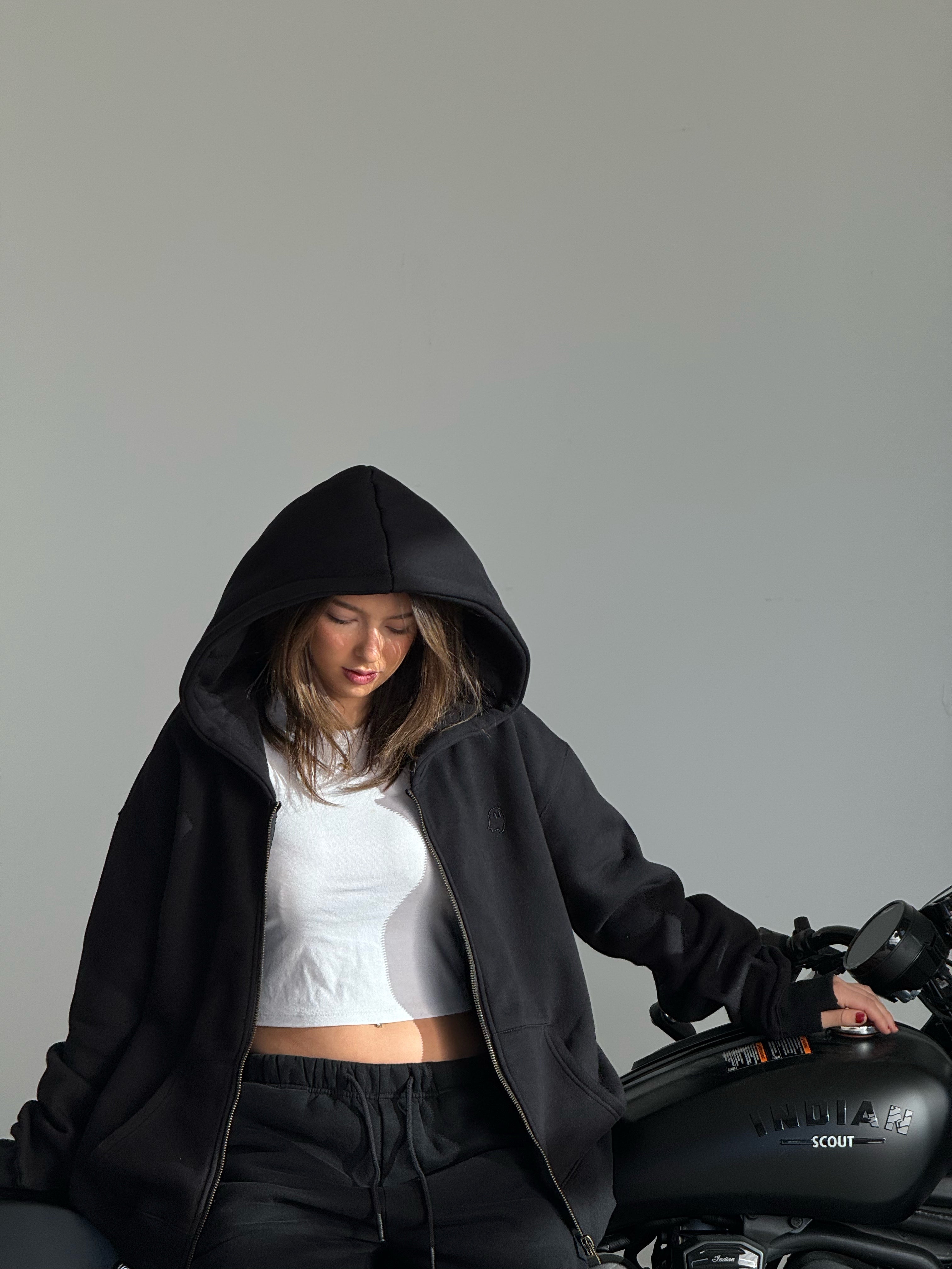 Person wearing a black hoodie and white top sitting next to a motorcycle on a gray background