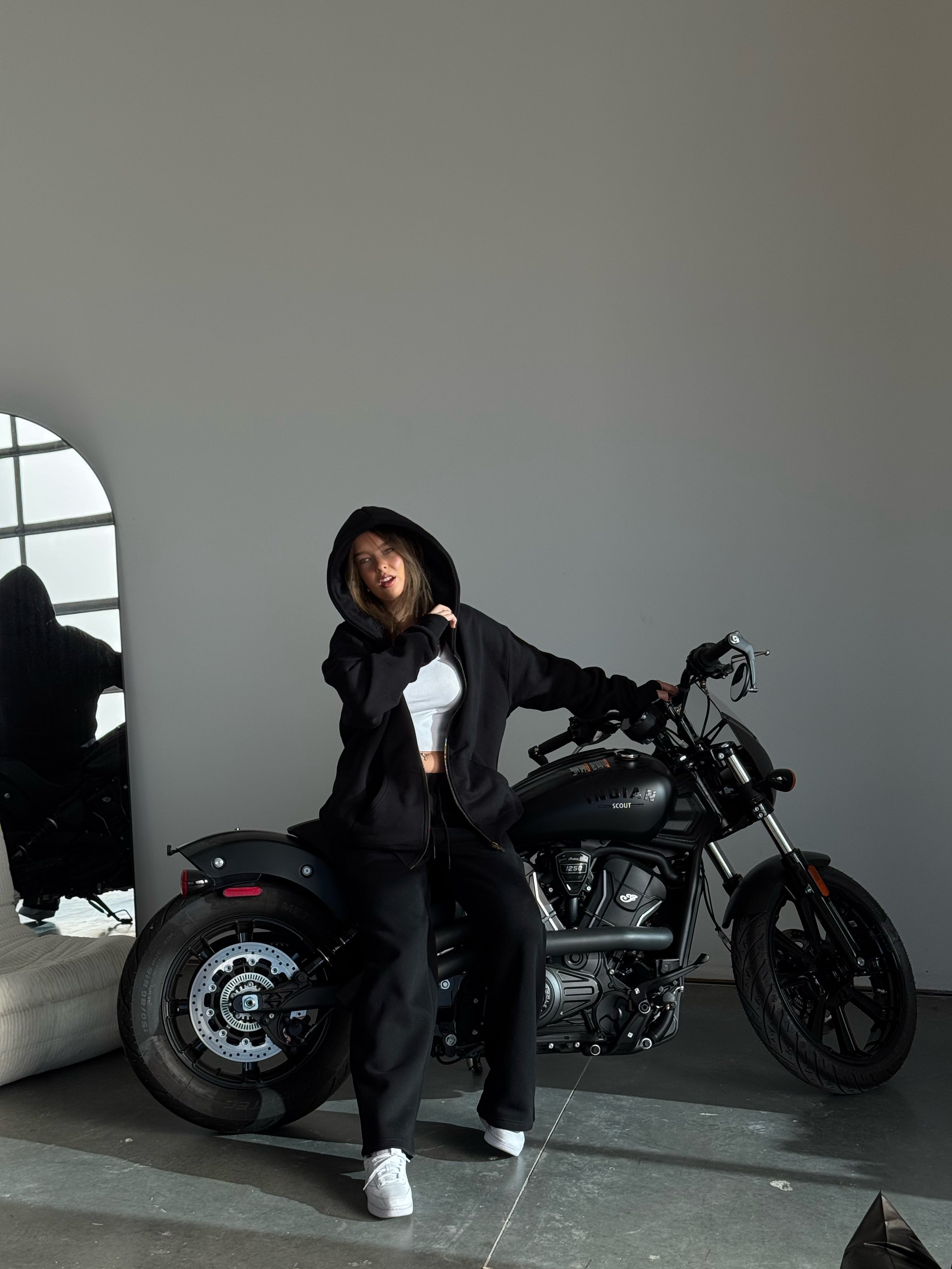 Person in black hoodie and pants standing next to a motorcycle in a minimalistic setting