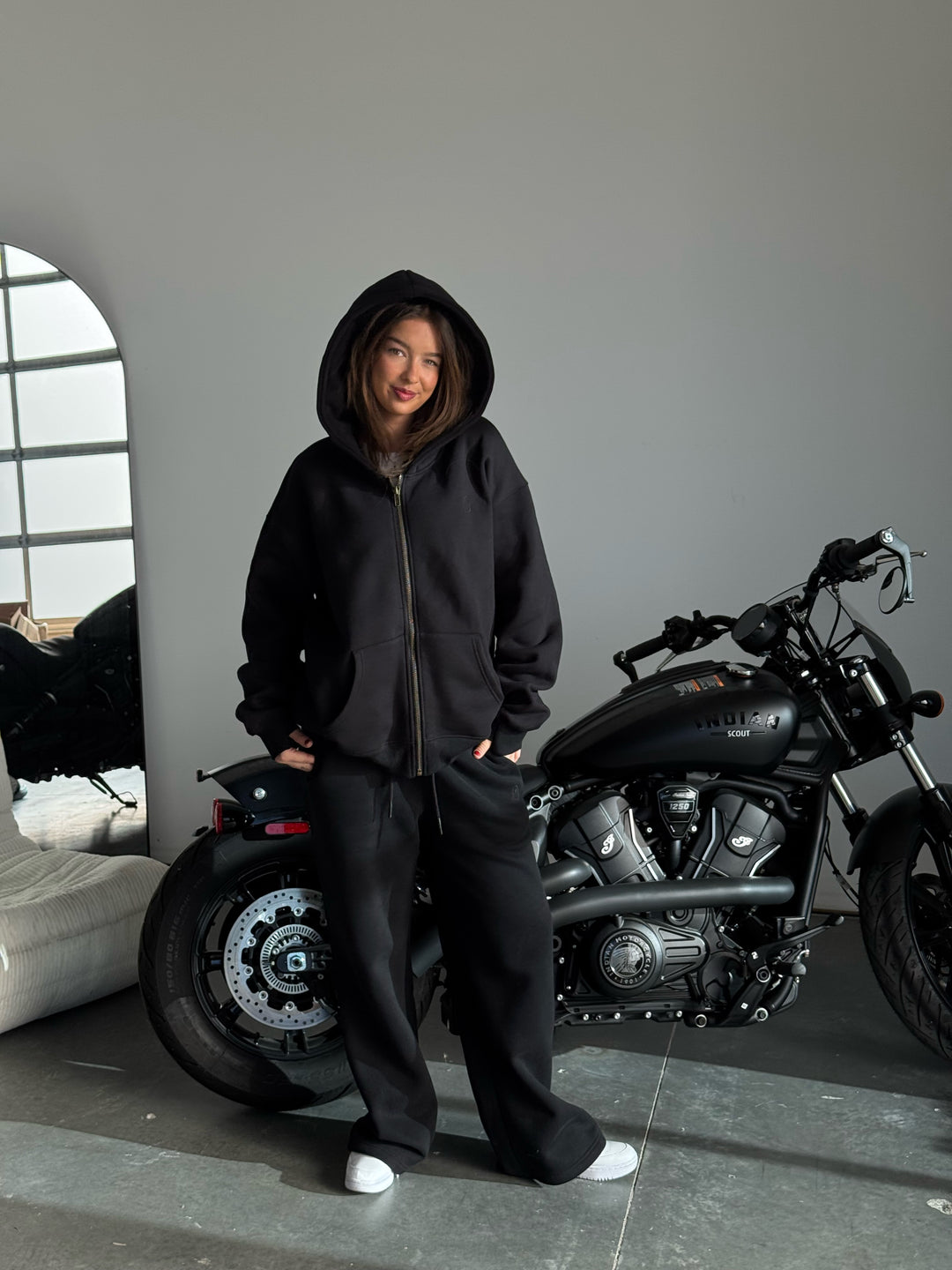 Person wearing a black hoodie and pants standing next to a motorcycle indoors.