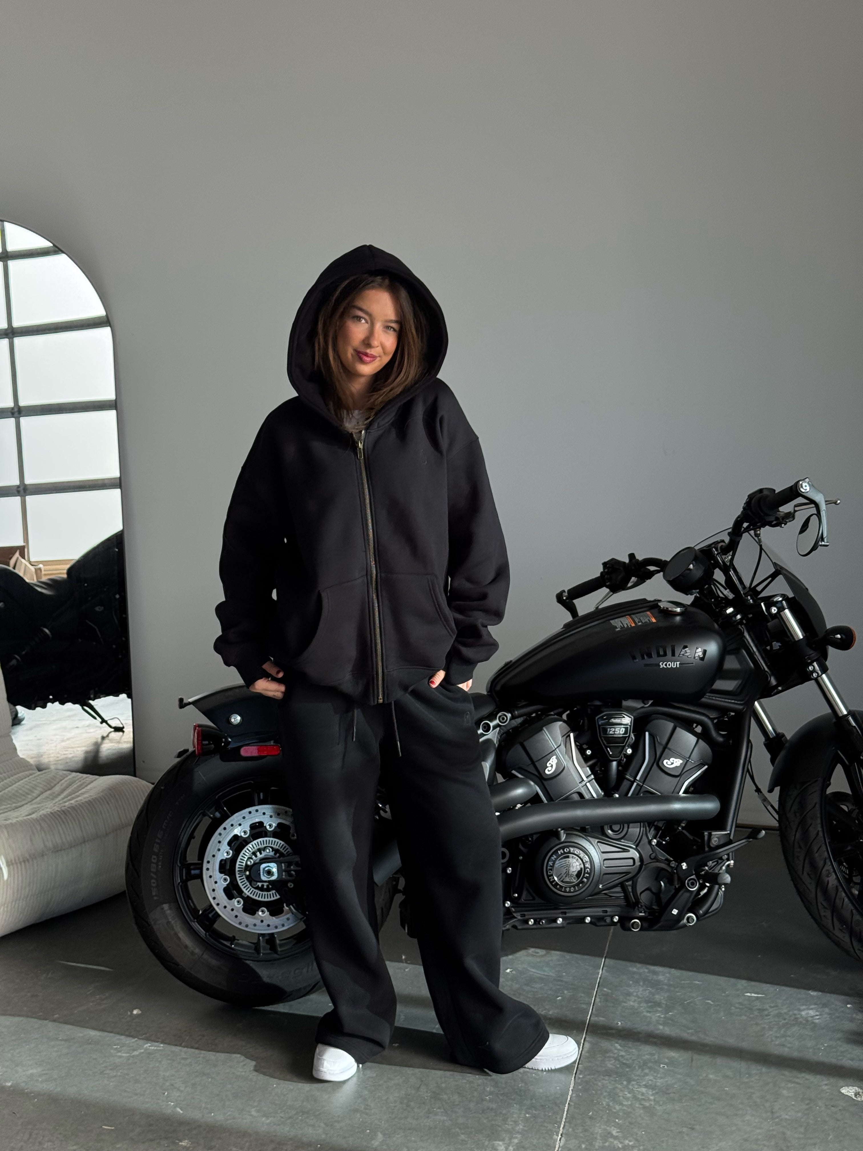 Person wearing a black hoodie and pants standing next to a motorcycle indoors.