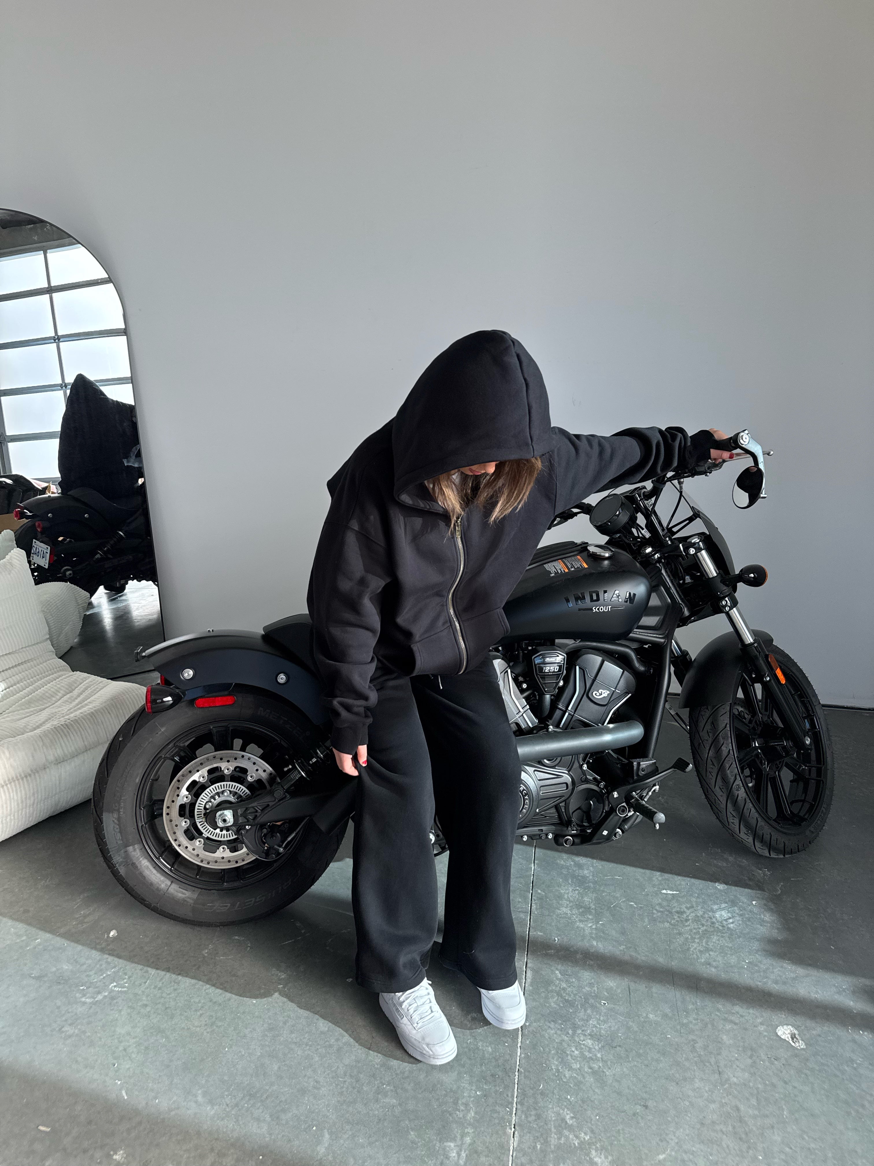 Person wearing a black hoodie and pants standing next to a black motorcycle indoors.