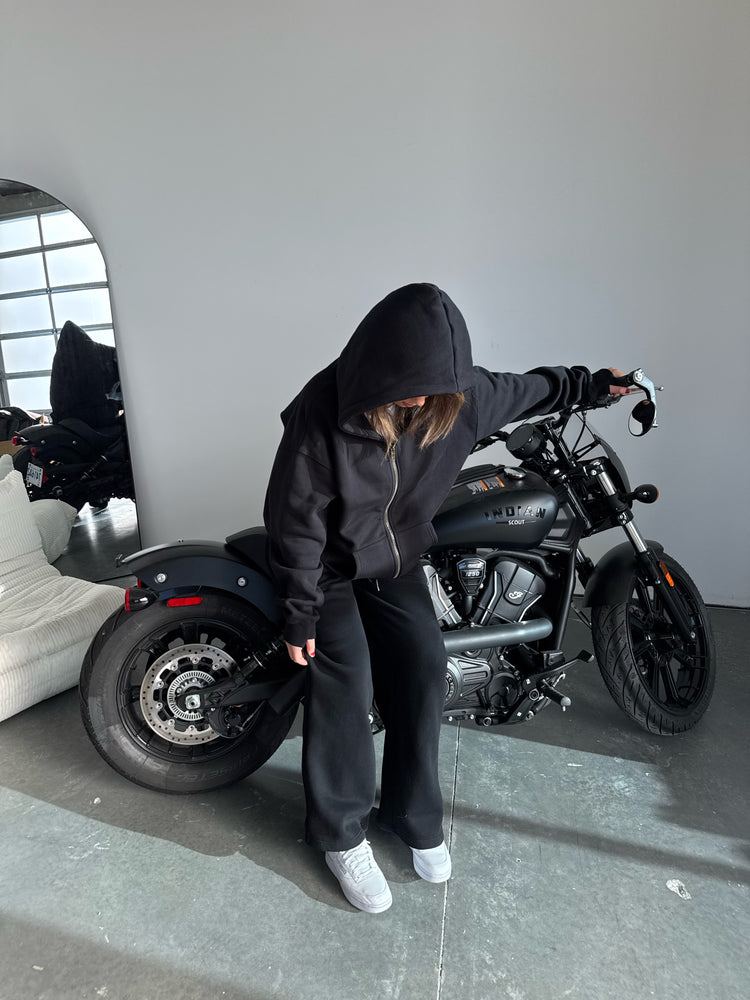 Person wearing a black hoodie and pants standing next to a black motorcycle indoors.