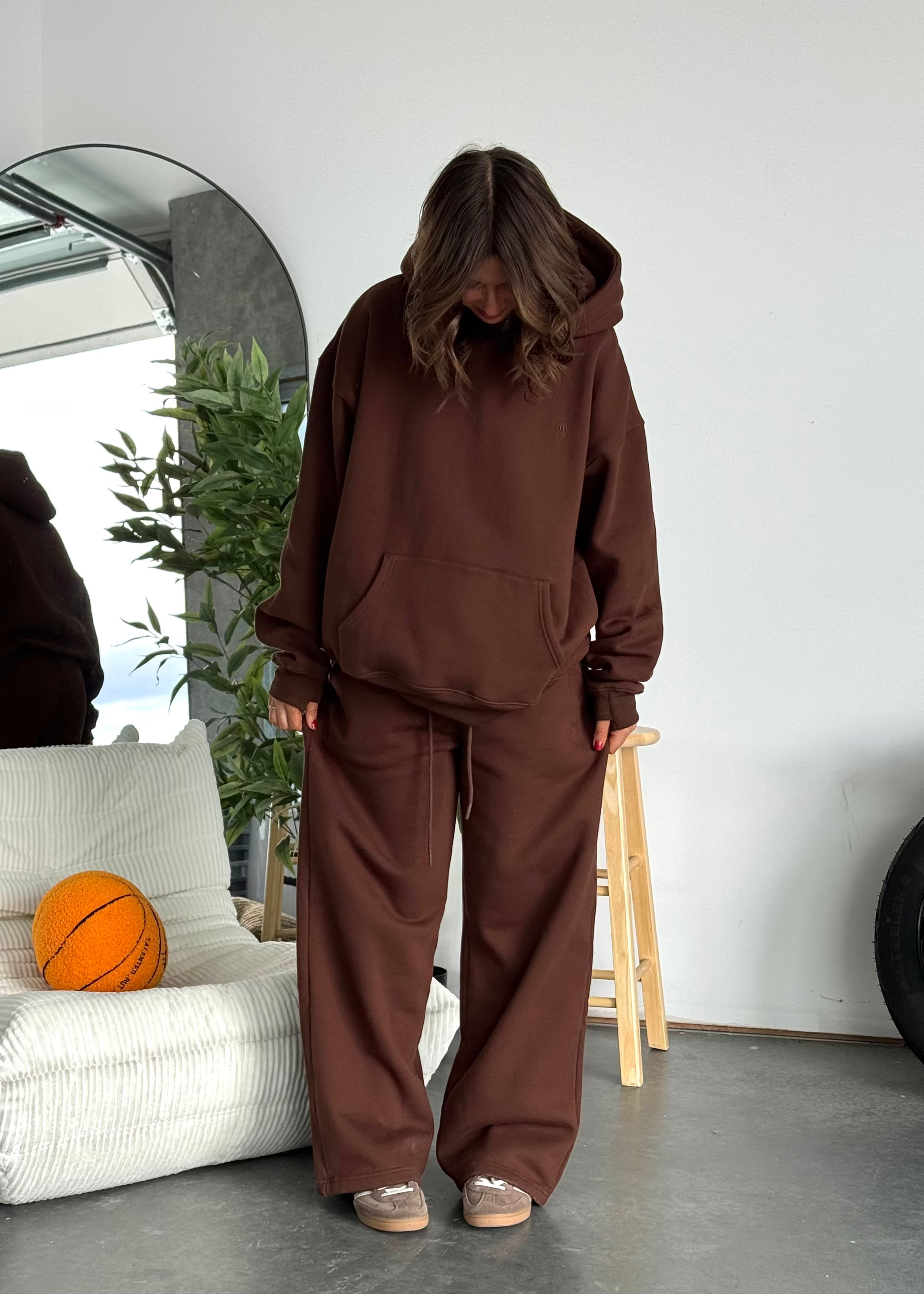 Person wearing a brown hoodie and pants standing in a room with a white wall, gray floor, and a plant.