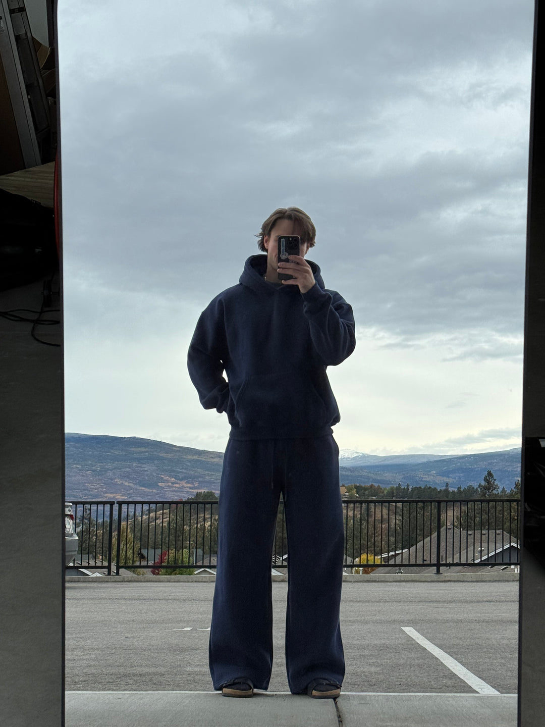 Person taking a mirror selfie wearing a dark blue tracksuit with a cloudy sky and mountains in the background.