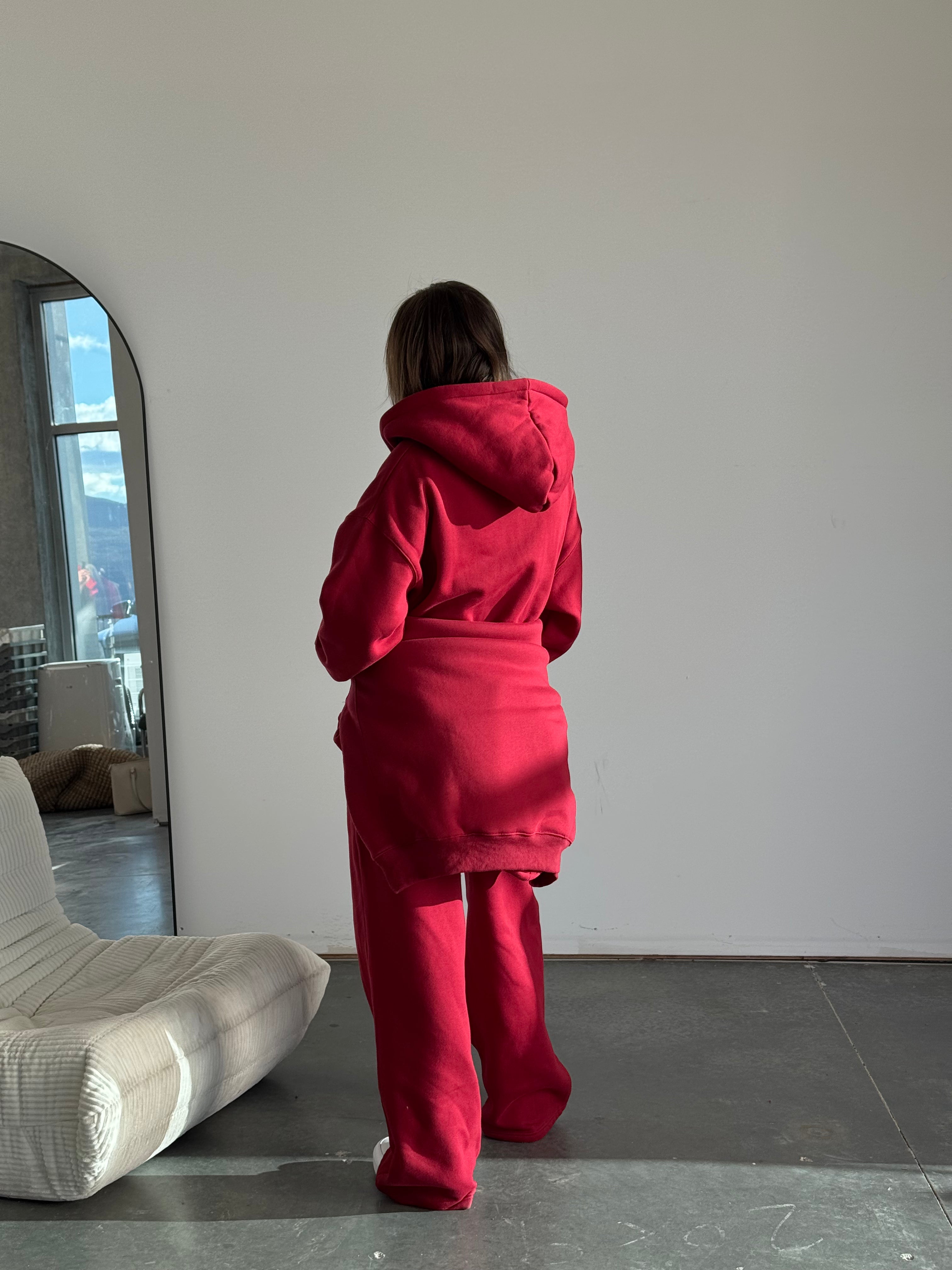 Person wearing a red hoodie and pants standing in a room with a mirror and chair.