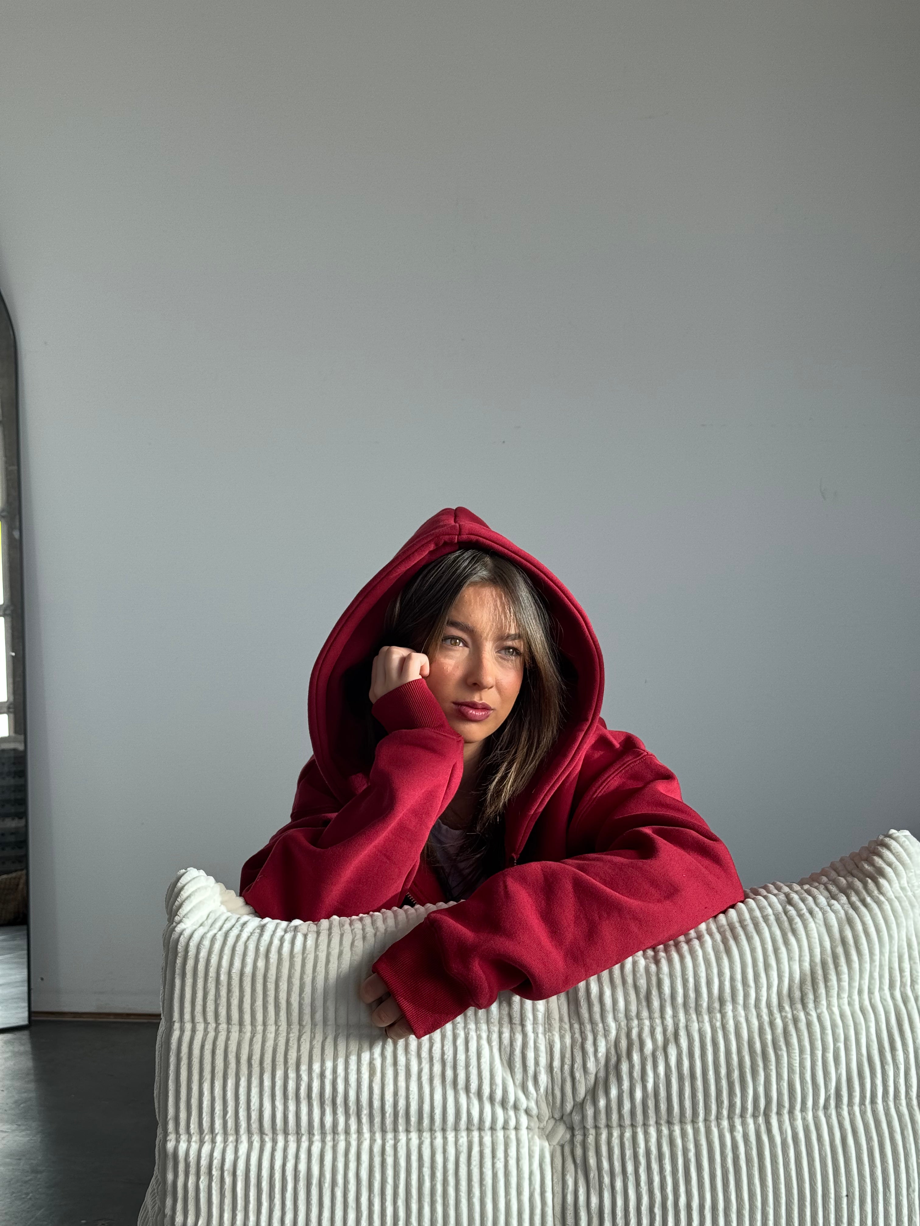 Person wearing a red hoodie sitting on a white couch against a plain wall.