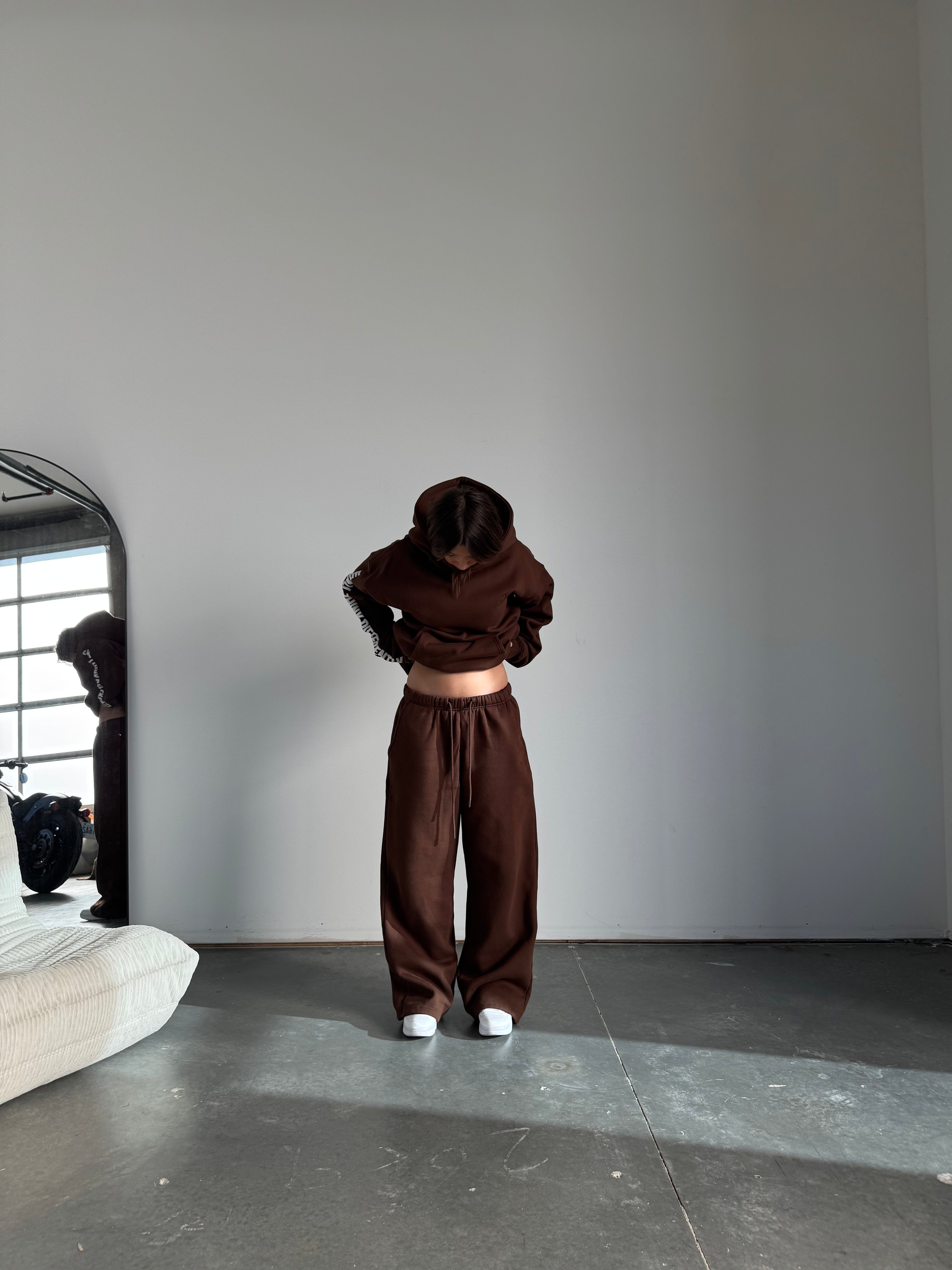 Person wearing a brown tracksuit standing in a minimalistic room with a mirror.