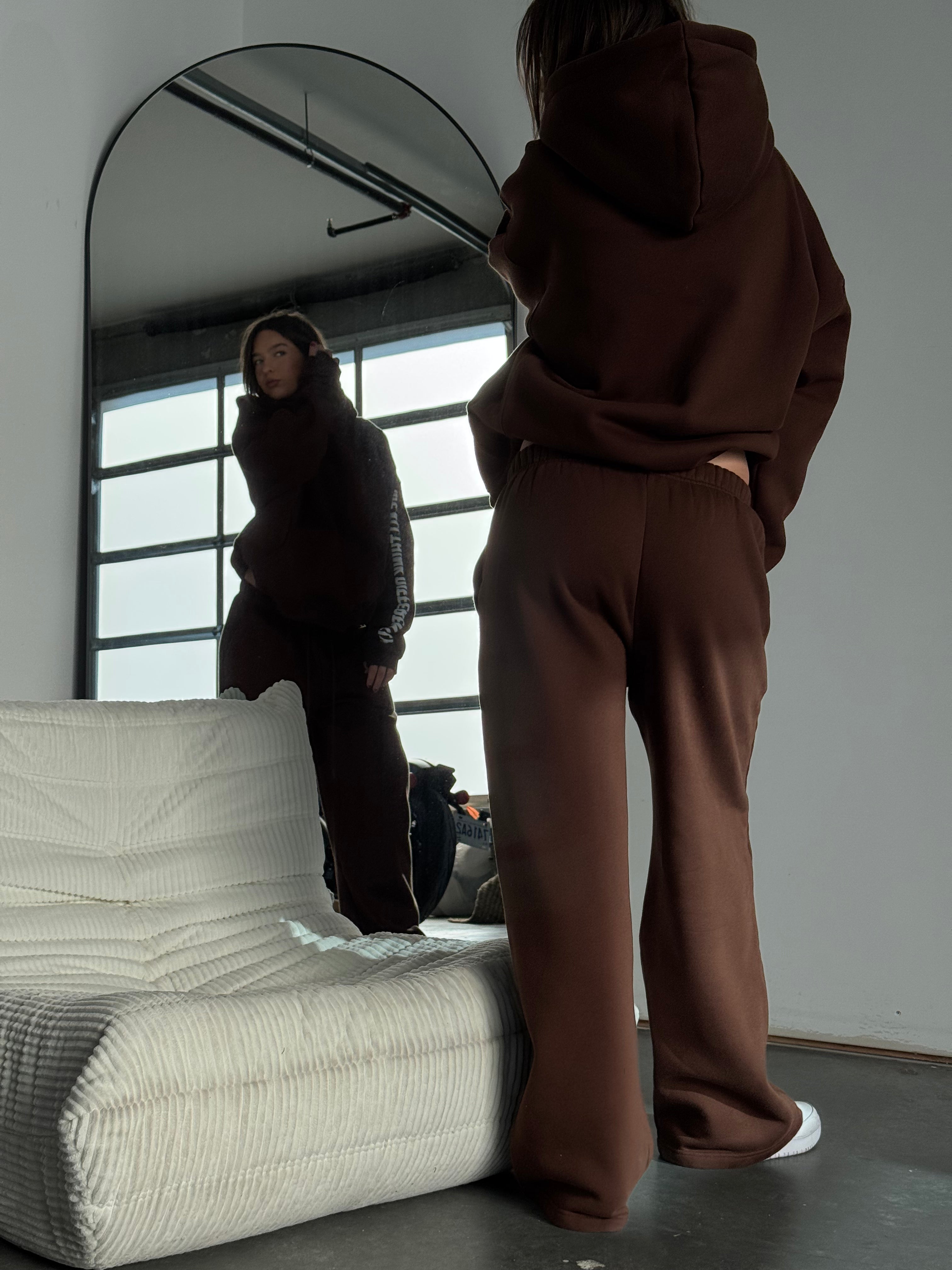 Person wearing a brown hoodie and pants standing in front of a mirror in a room with a white sofa.