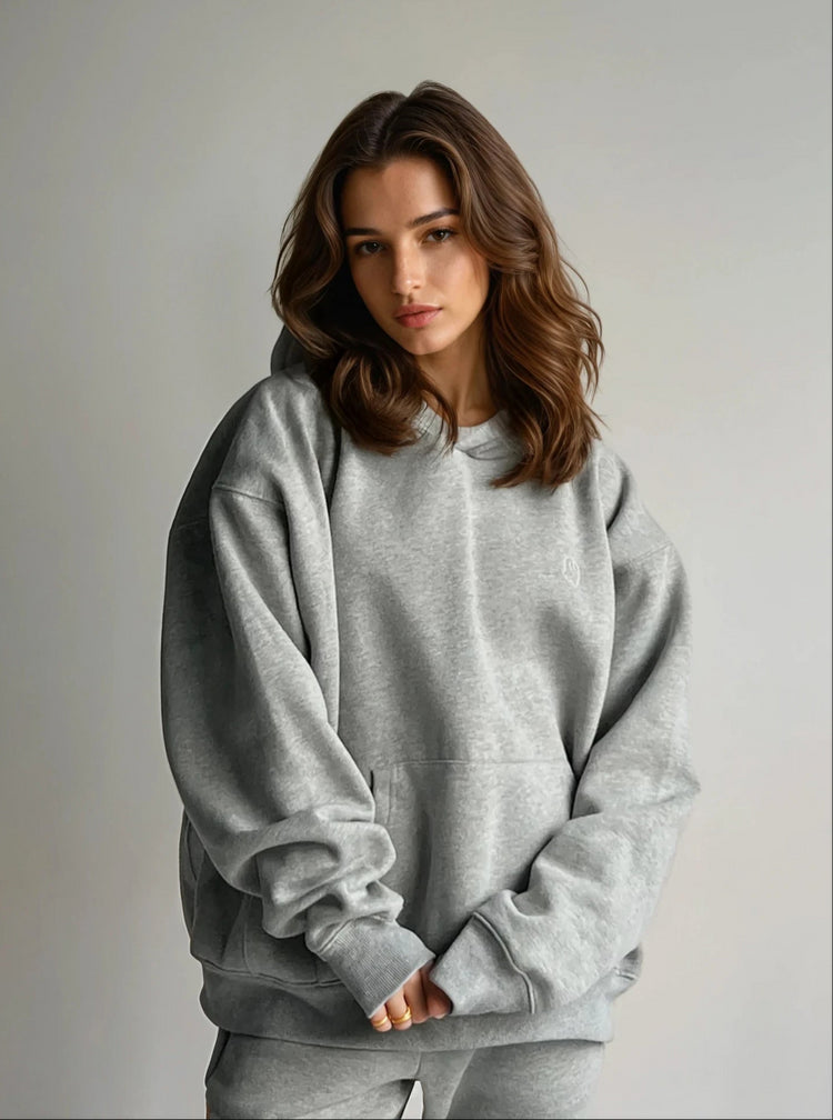 A person wearing a loose-fitting grey hoodie with a hood, standing against a grey background.