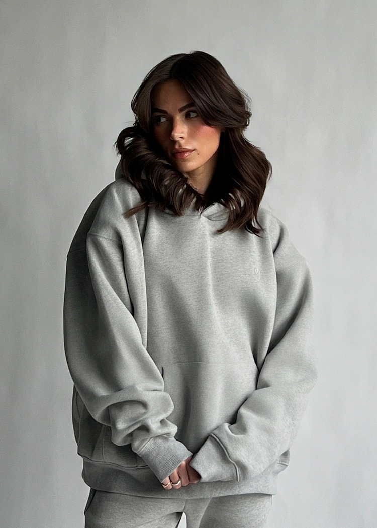 A woman with wavy brown hair wearing an oversized light gray heather sweatshirt and matching sweatpants, standing against a neutral gray studio background.