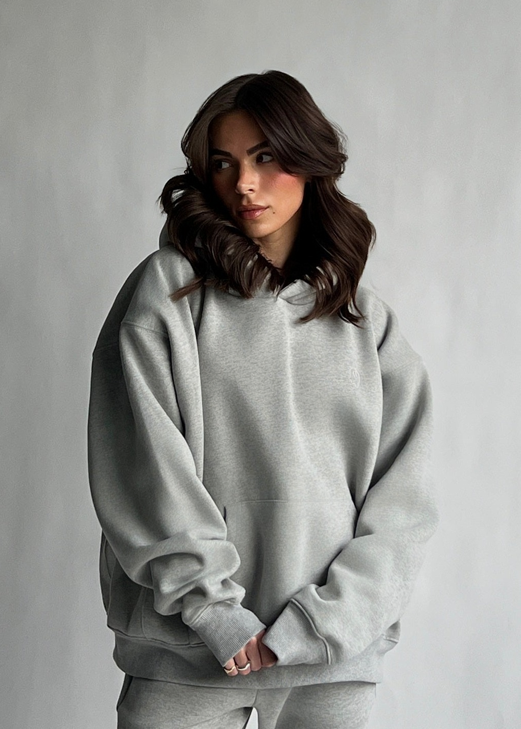 A woman with wavy brown hair wearing an oversized light gray heather sweatshirt and matching sweatpants, standing against a neutral gray studio background.