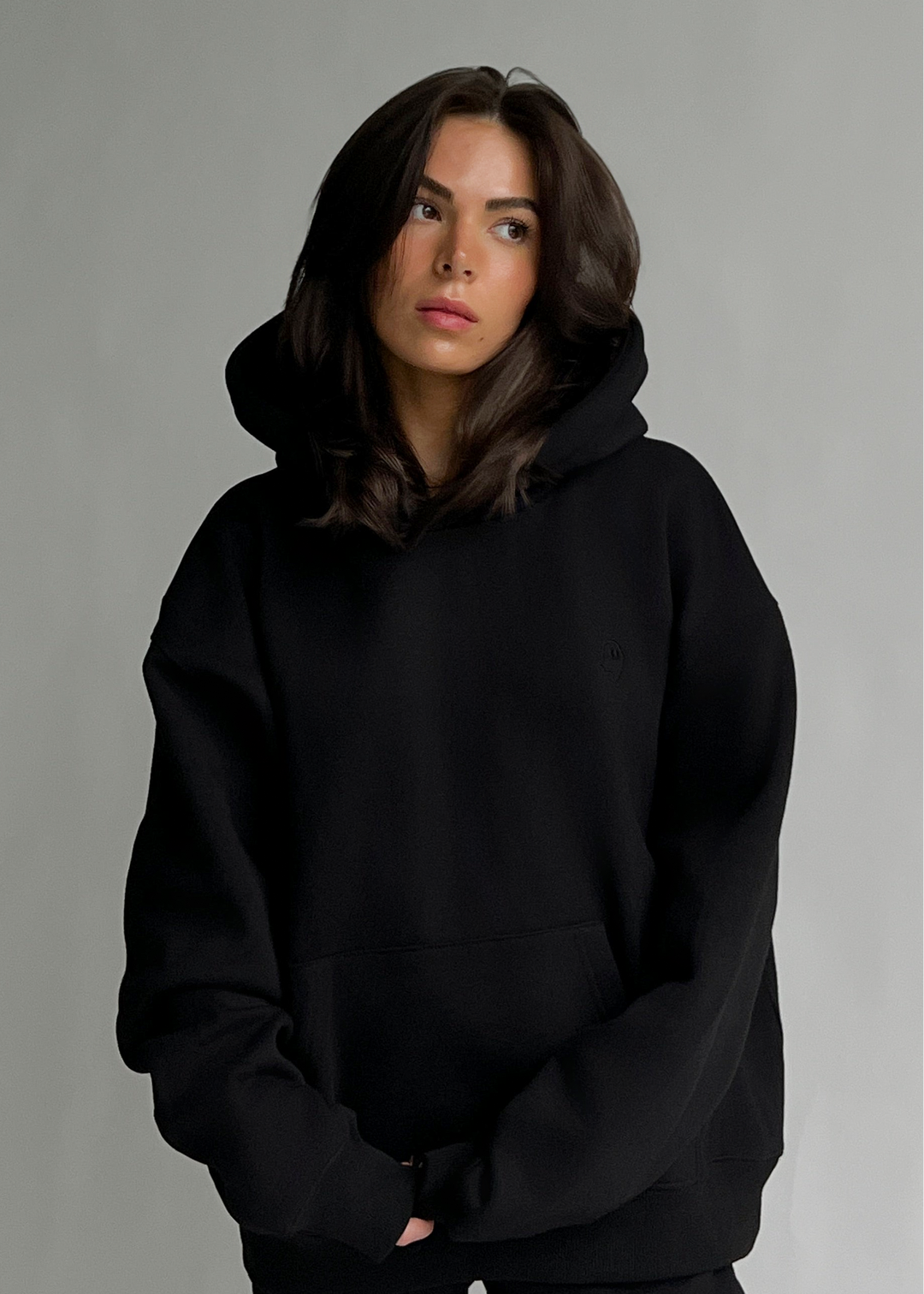 A woman wearing an oversized black pullover hoodie with a front kangaroo pocket, photographed against a plain light gray studio background.