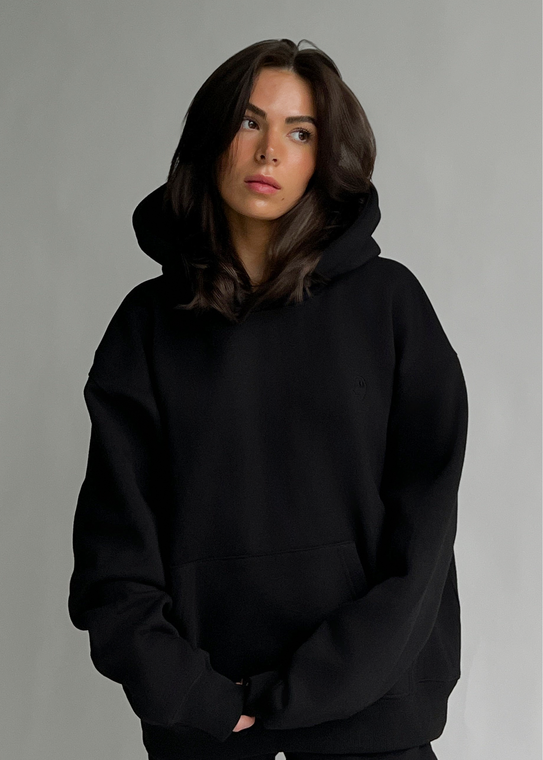 A woman wearing an oversized black pullover hoodie with a front kangaroo pocket, photographed against a plain light gray studio background.