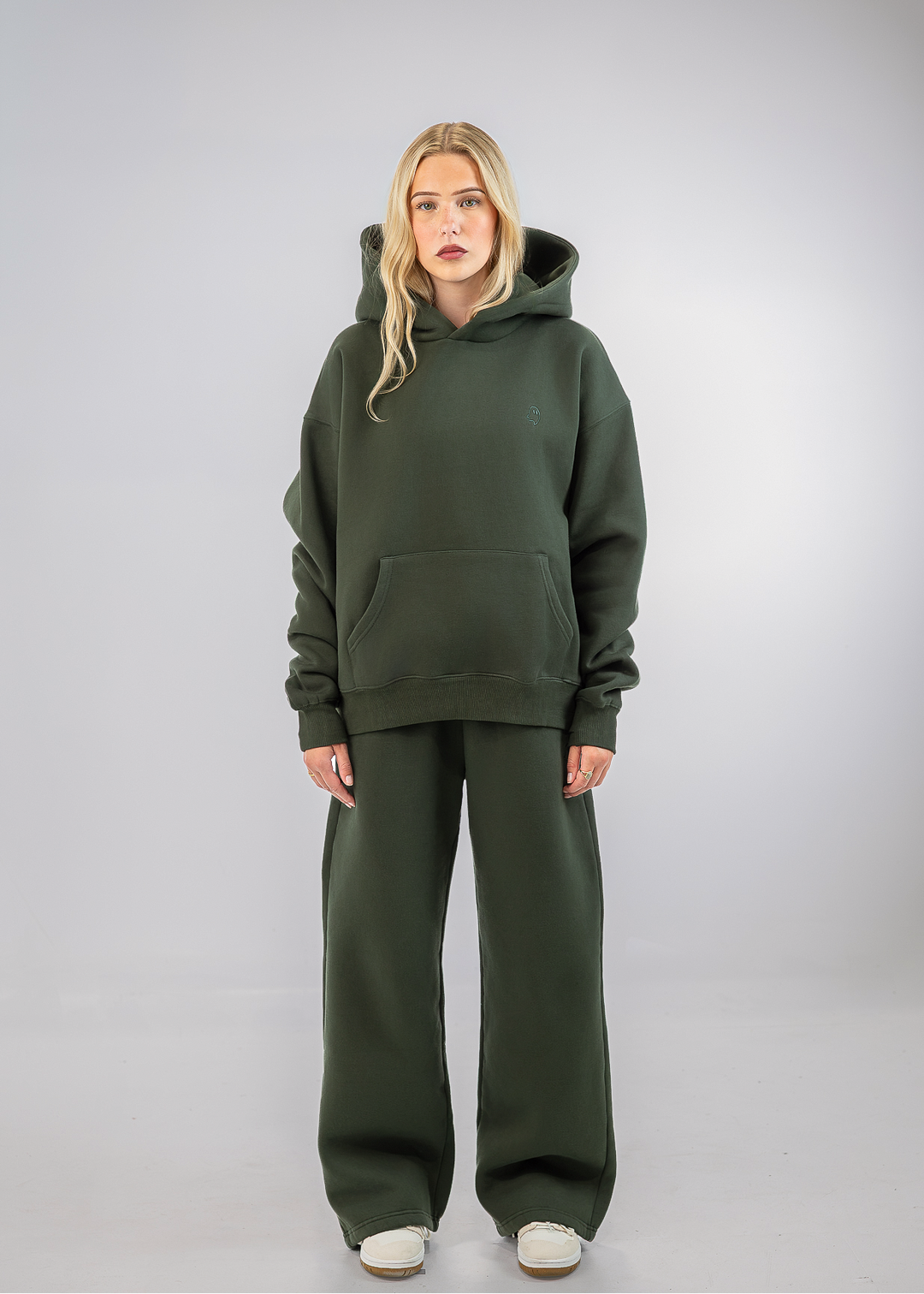 A blonde model stands against a light gray studio background wearing an oversized dark olive green hoodie and matching wide-leg sweatpants with white sneakers.
