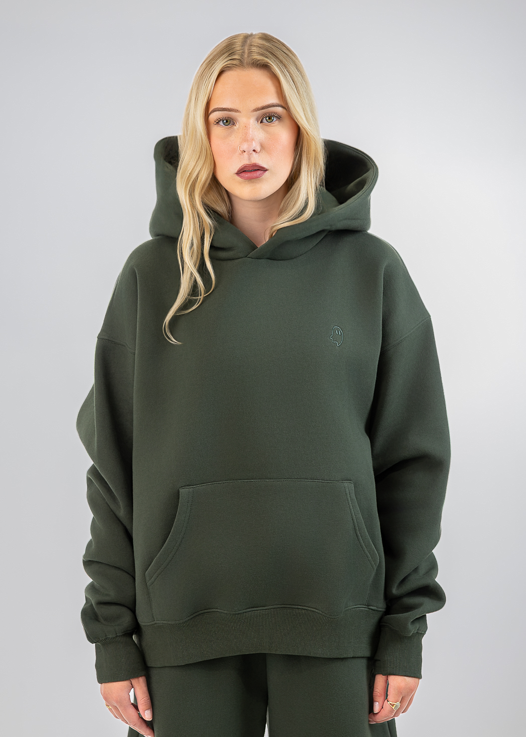 A blonde woman modeling an oversized dark olive green hoodie with a matching pair of sweatpants against a light gray studio background.