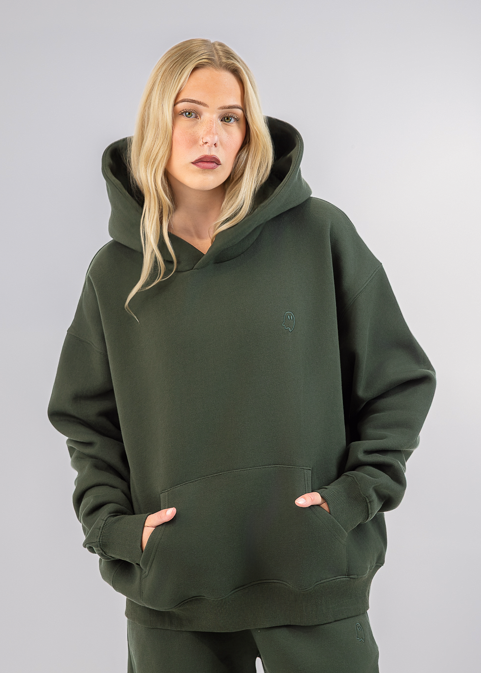 A blonde woman wearing an oversized dark olive green hoodie and matching sweatpants, standing against a neutral light gray studio background.