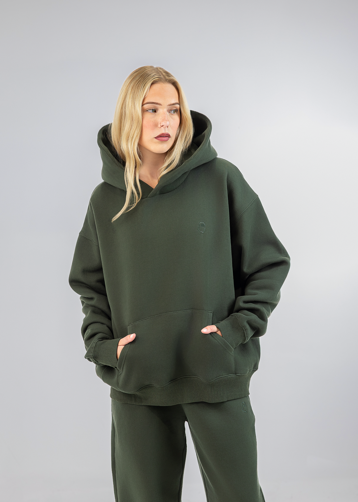 A blonde woman wearing an oversized dark olive green hoodie and matching sweatpants, standing against a neutral gray studio background.