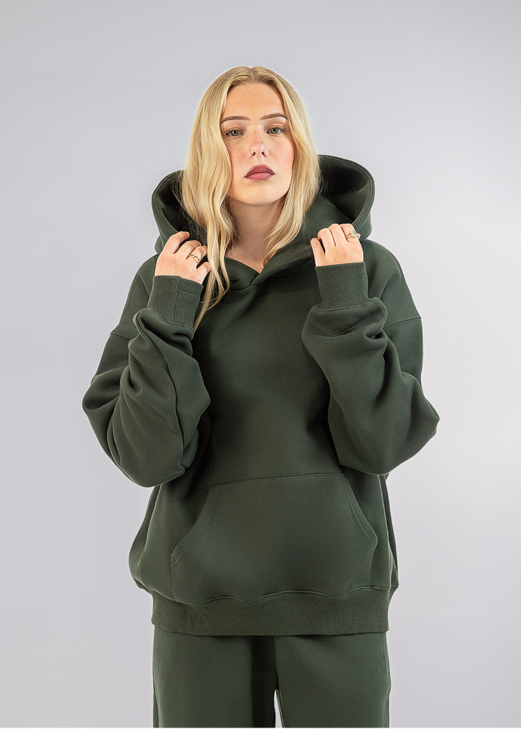 A blonde woman wearing an olive green pullover hoodie and matching sweatpants, standing against a neutral gray studio background.