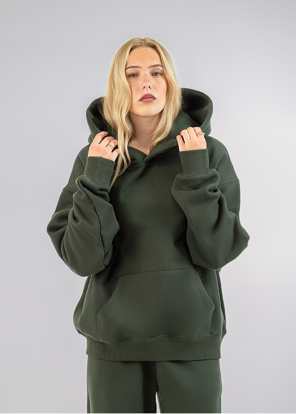 A blonde woman wearing an olive green pullover hoodie and matching sweatpants, standing against a neutral gray studio background.