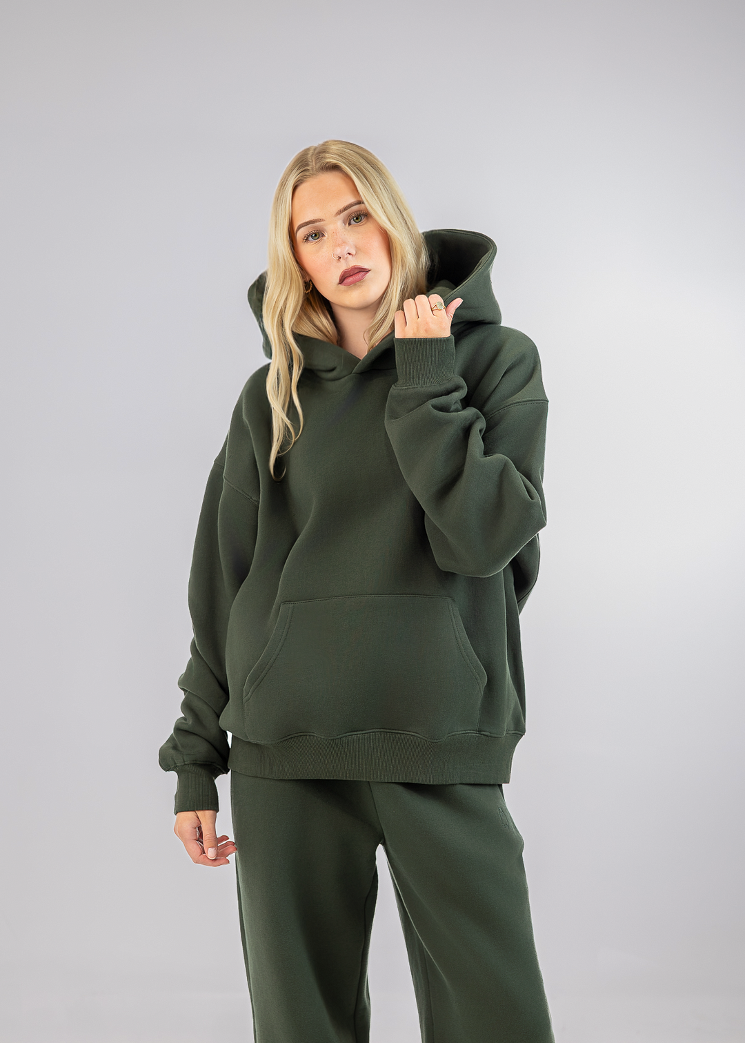 A blonde woman modeling an oversized dark green hoodie and matching sweatpants against a light gray studio background.