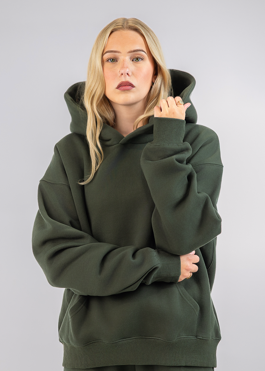A woman modeling an oversized dark olive green hoodie and matching sweatpants against a light gray studio background.