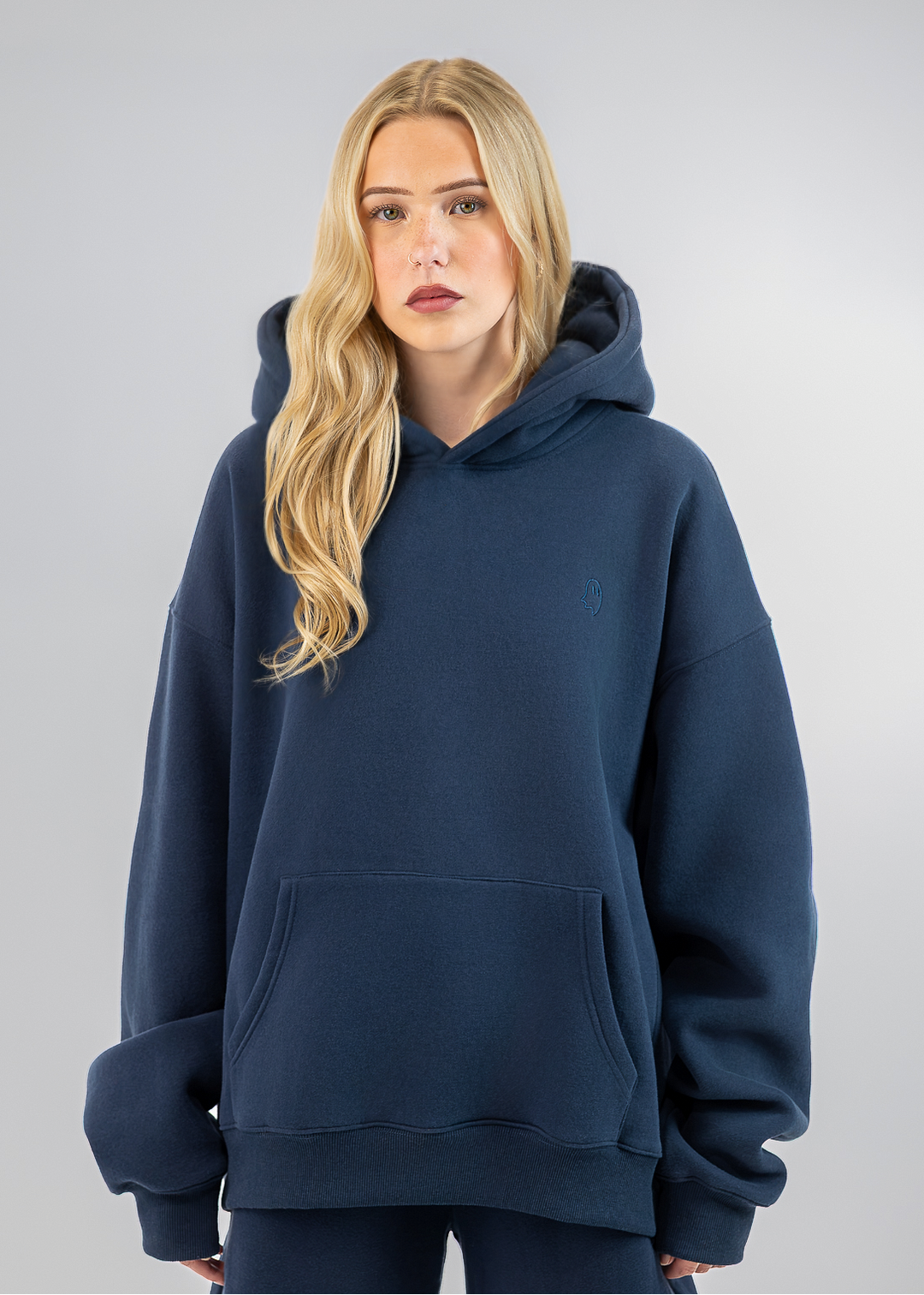 A blonde woman wearing an oversized navy blue hoodie and matching sweatpants, standing against a neutral gray studio background.
