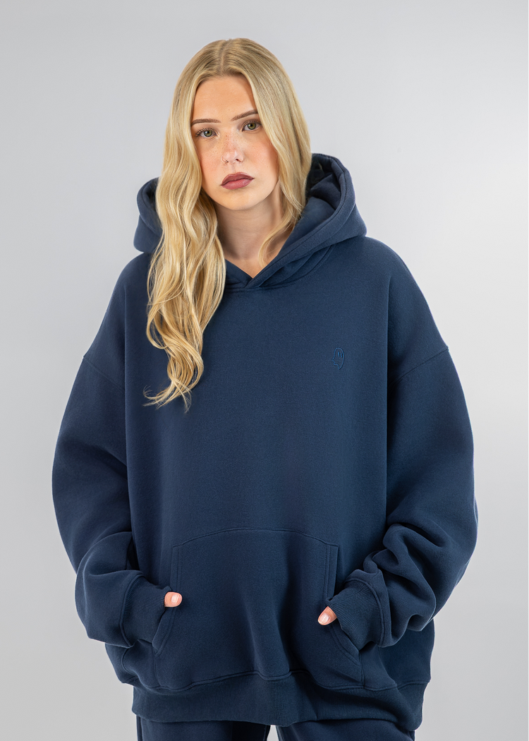 A blonde woman wearing an oversized navy blue hoodie with a small embroidered polo player logo on the left chest, paired with matching navy pants, set against a solid light gray studio background.