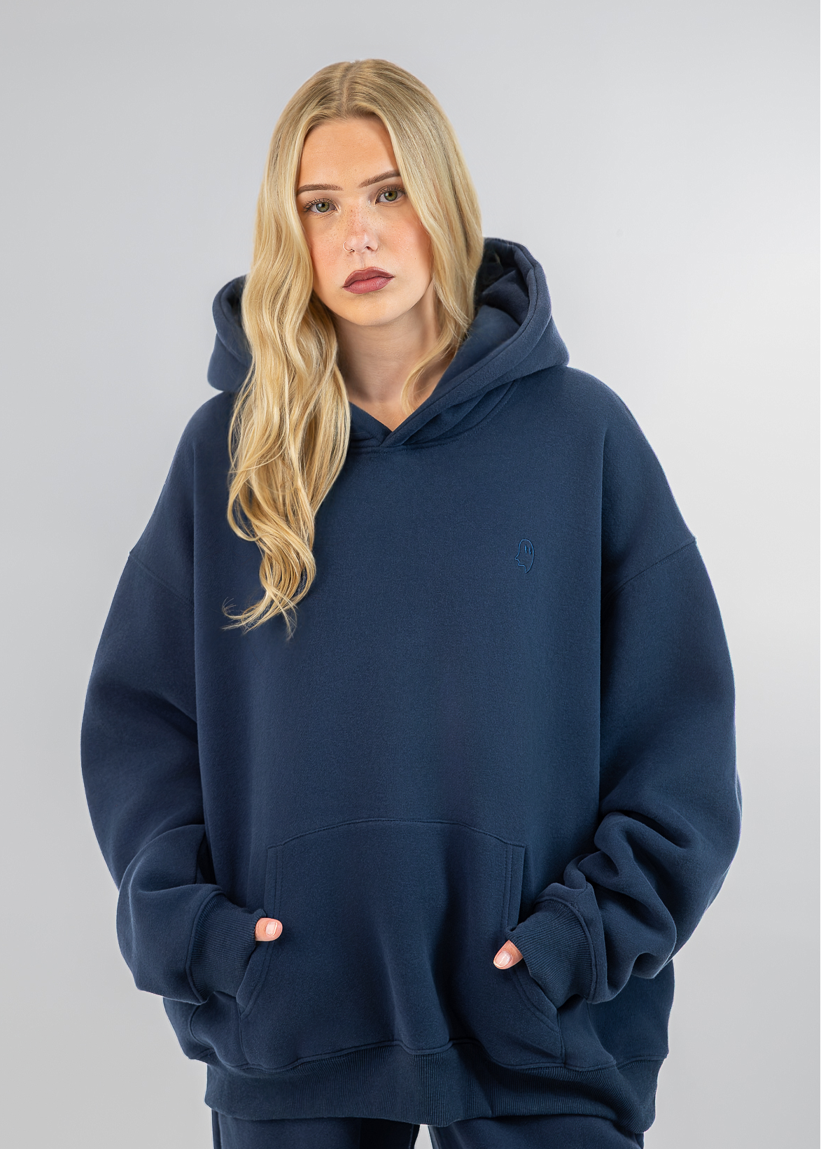 A blonde woman wearing an oversized navy blue hoodie with a small embroidered polo player logo on the left chest, paired with matching navy pants, set against a solid light gray studio background.
