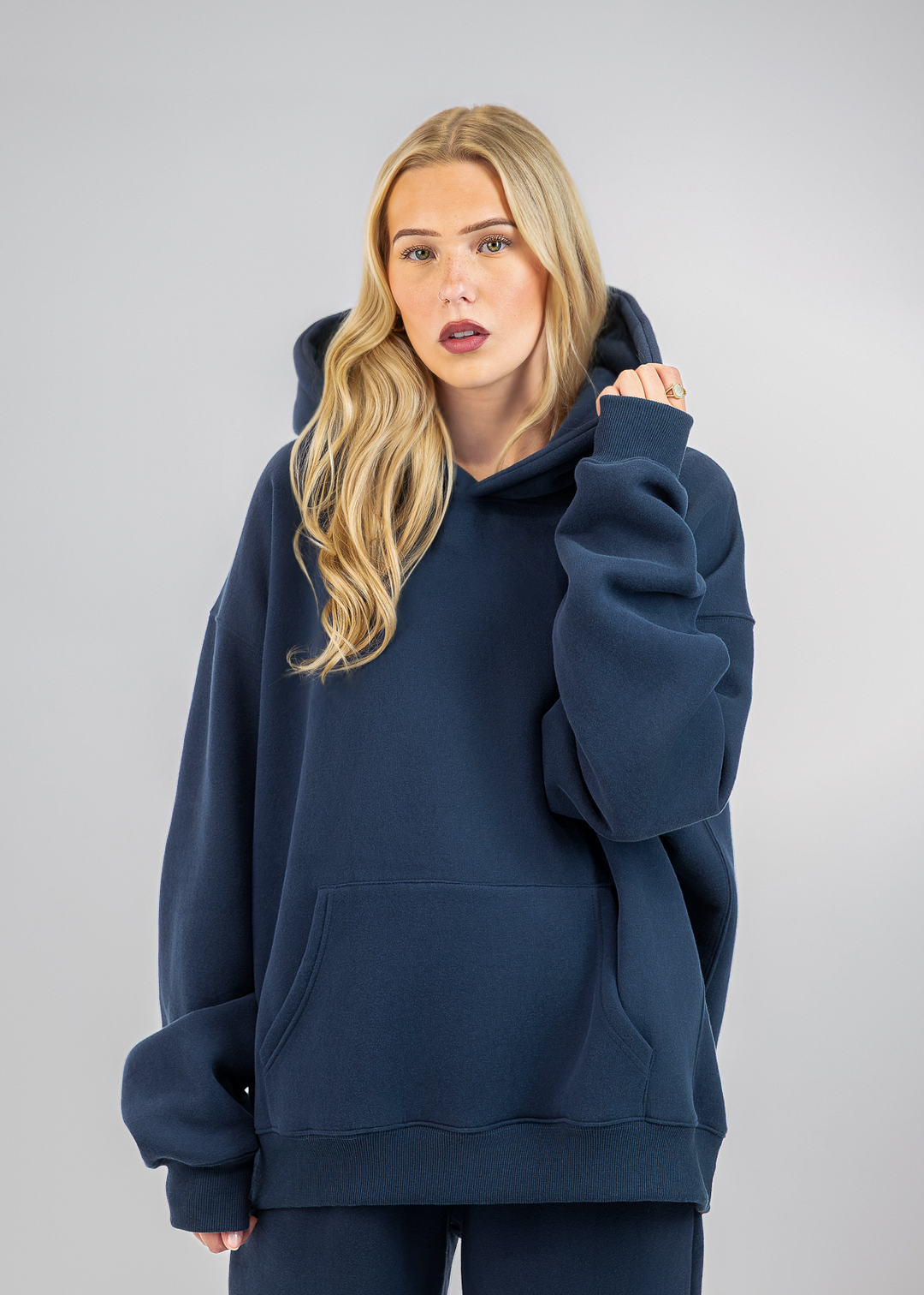 A blonde woman wearing an oversized navy blue hoodie and matching sweatpants, standing against a neutral gray studio background.