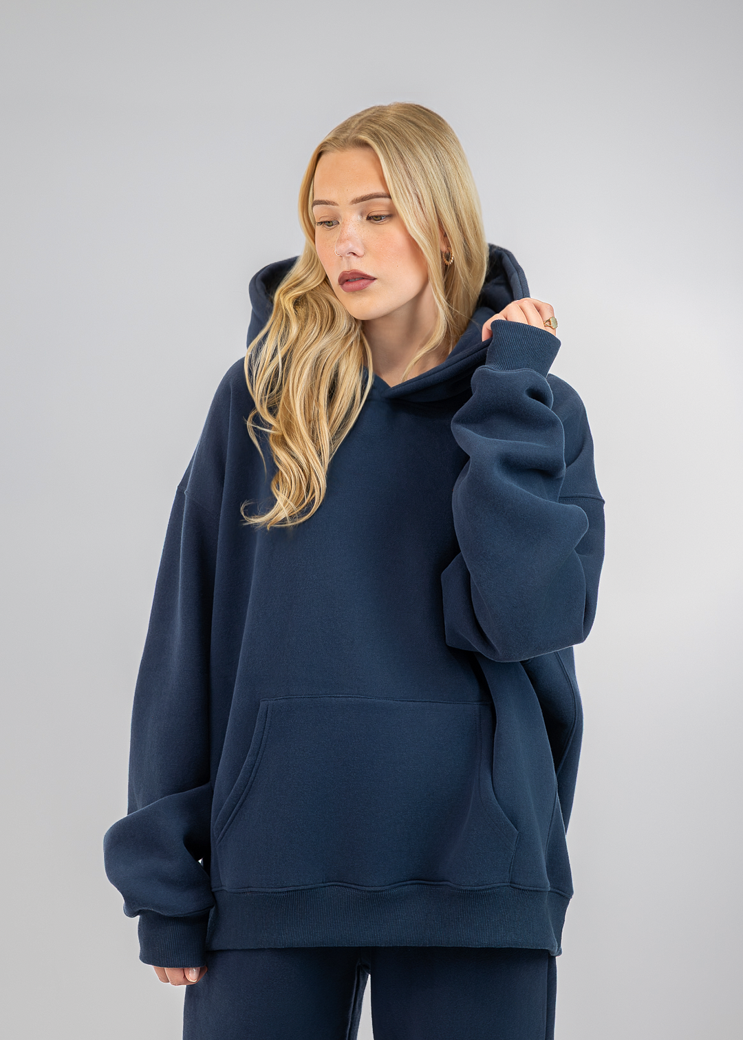 A blonde woman wearing an oversized navy blue hoodie and matching sweatpants, standing against a neutral light gray studio background.