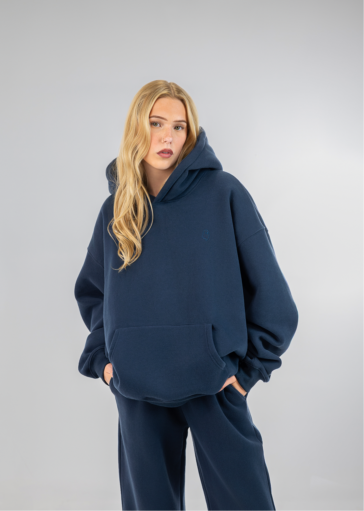 A blonde woman wearing an oversized navy blue hoodie and matching sweatpants, standing against a light gray studio background.
