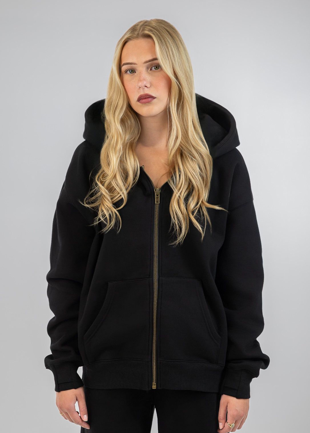 A blonde woman wearing a black zip-up hoodie with a gold zipper and black pants, standing against a light gray studio background.