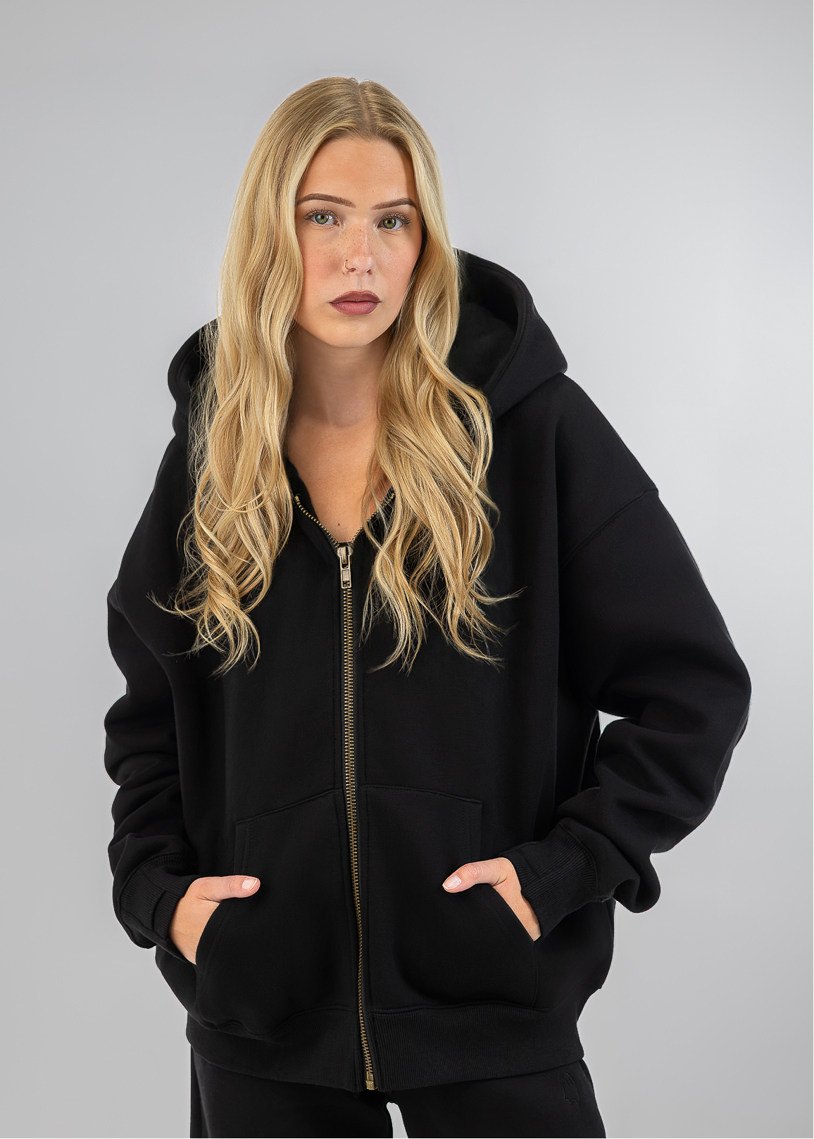 A blonde woman wearing a black zip-up hoodie with a gold zipper and black pants, standing against a light gray studio background.