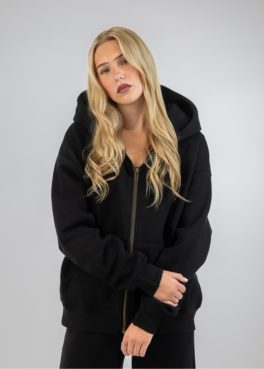 A blonde woman wearing a black zip-up hoodie with a gold zipper and black sweatpants, posing against a light gray studio background.