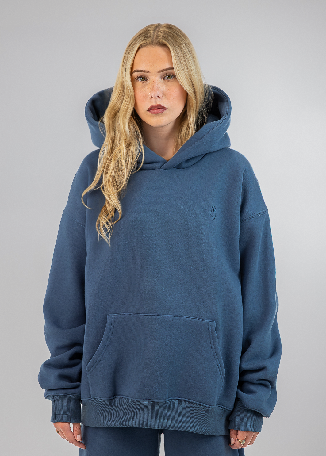 A blonde woman wearing an oversized dusty blue hoodie with a kangaroo pocket and matching sweatpants, standing against a neutral gray studio background.
