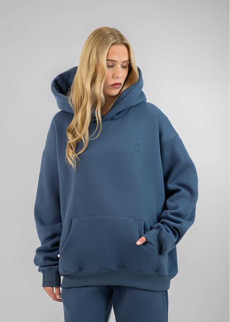 A woman wearing an oversized slate blue hoodie and matching sweatpants, standing against a neutral gray studio background.