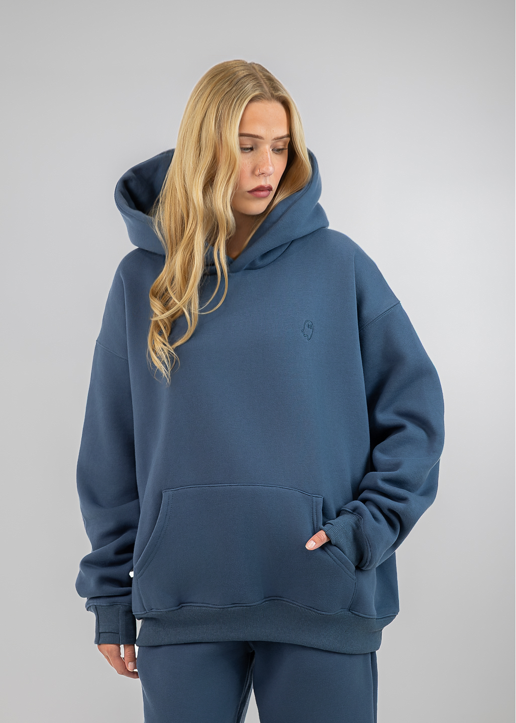 A woman wearing an oversized slate blue hoodie and matching sweatpants, standing against a neutral gray studio background.