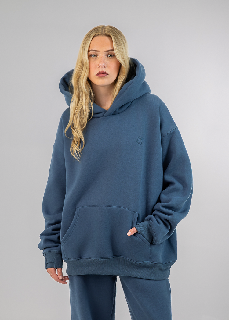 A blonde woman modeling an oversized slate blue hoodie and matching sweatpants against a neutral gray studio background.