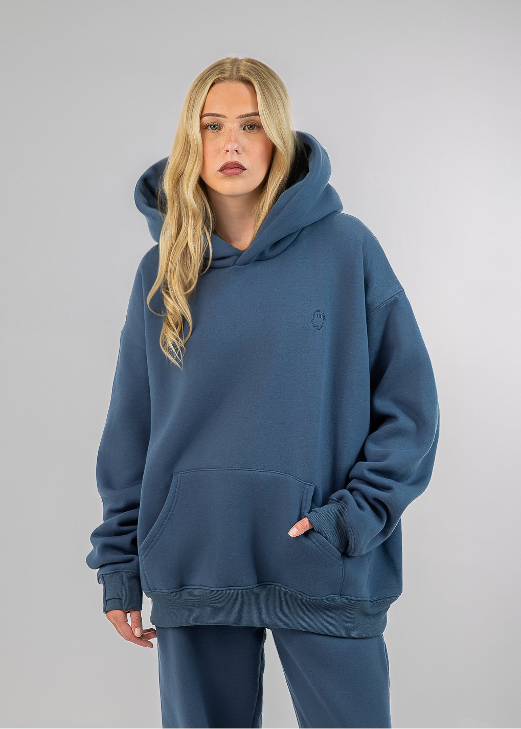 A blonde woman modeling an oversized slate blue hoodie and matching sweatpants against a neutral gray studio background.