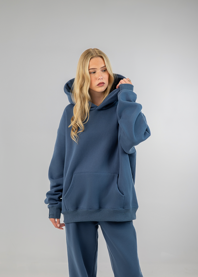 A woman modeling an oversized slate blue hoodie and matching sweatpants against a neutral gray studio background.