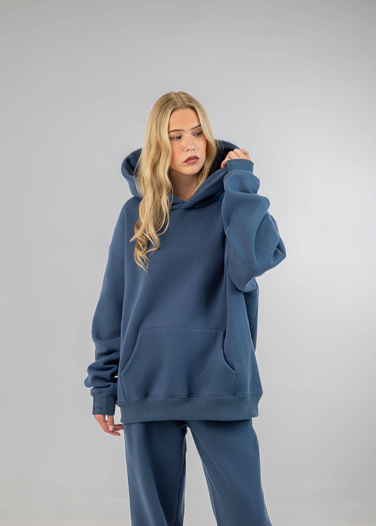 A woman modeling an oversized slate blue hoodie and matching sweatpants against a neutral gray studio background.