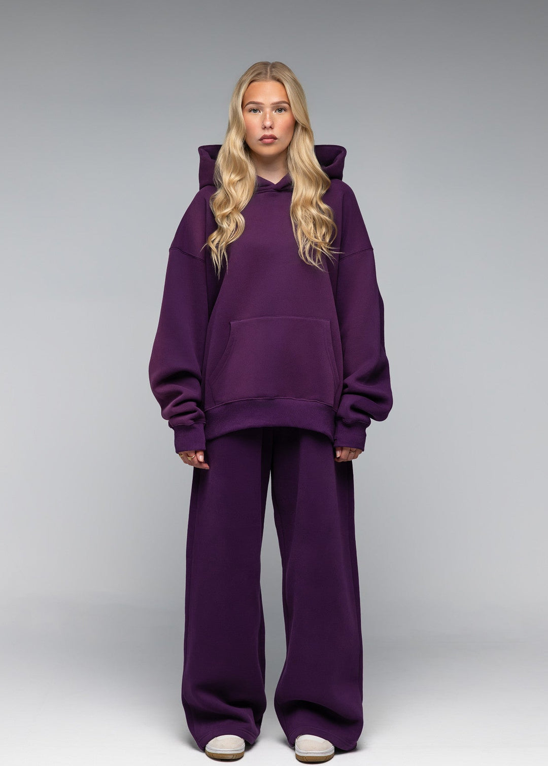 A blonde model stands against a gray studio background wearing an oversized matching purple hoodie and wide-leg sweatpants set.