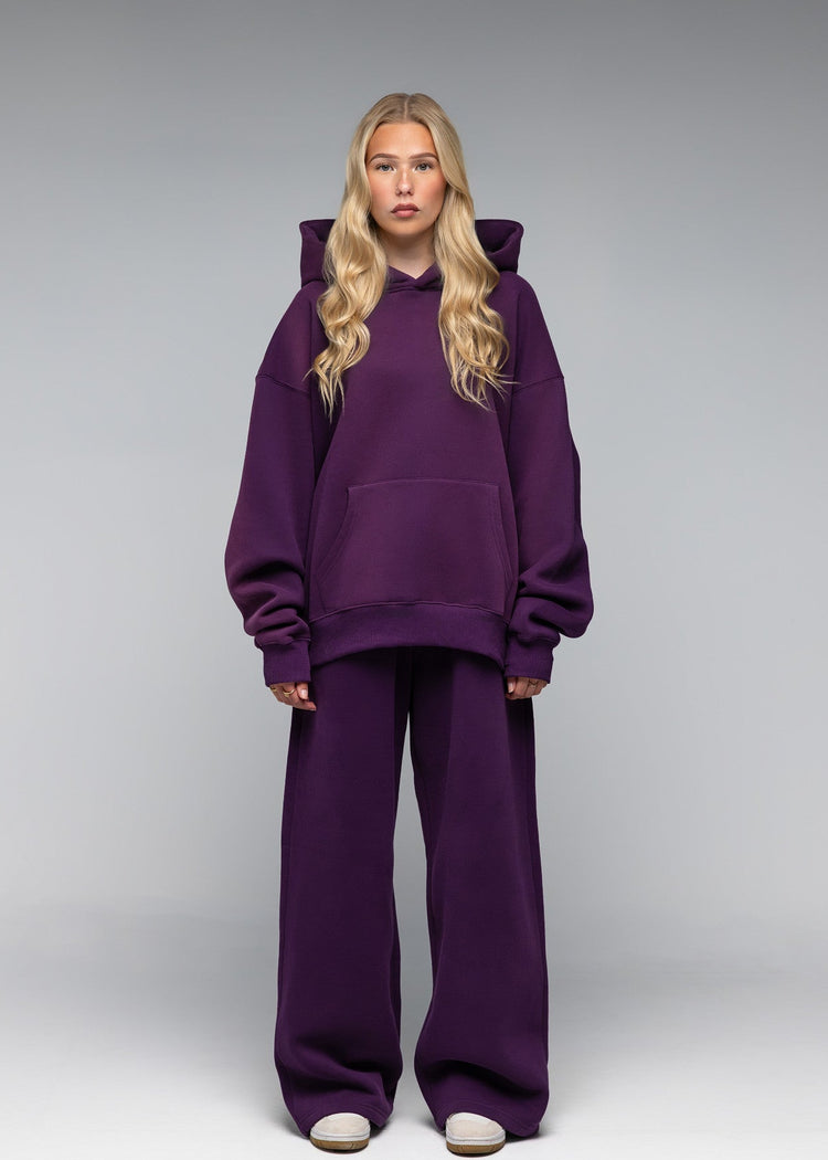 A blonde model stands against a gray studio background wearing an oversized matching purple hoodie and wide-leg sweatpants set.