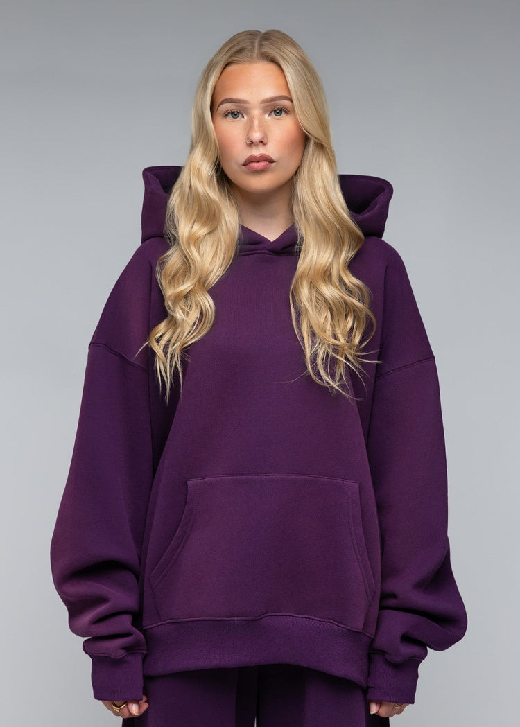 A blonde woman modeling an oversized deep purple hoodie and matching wide-leg sweatpants against a neutral gray studio background.