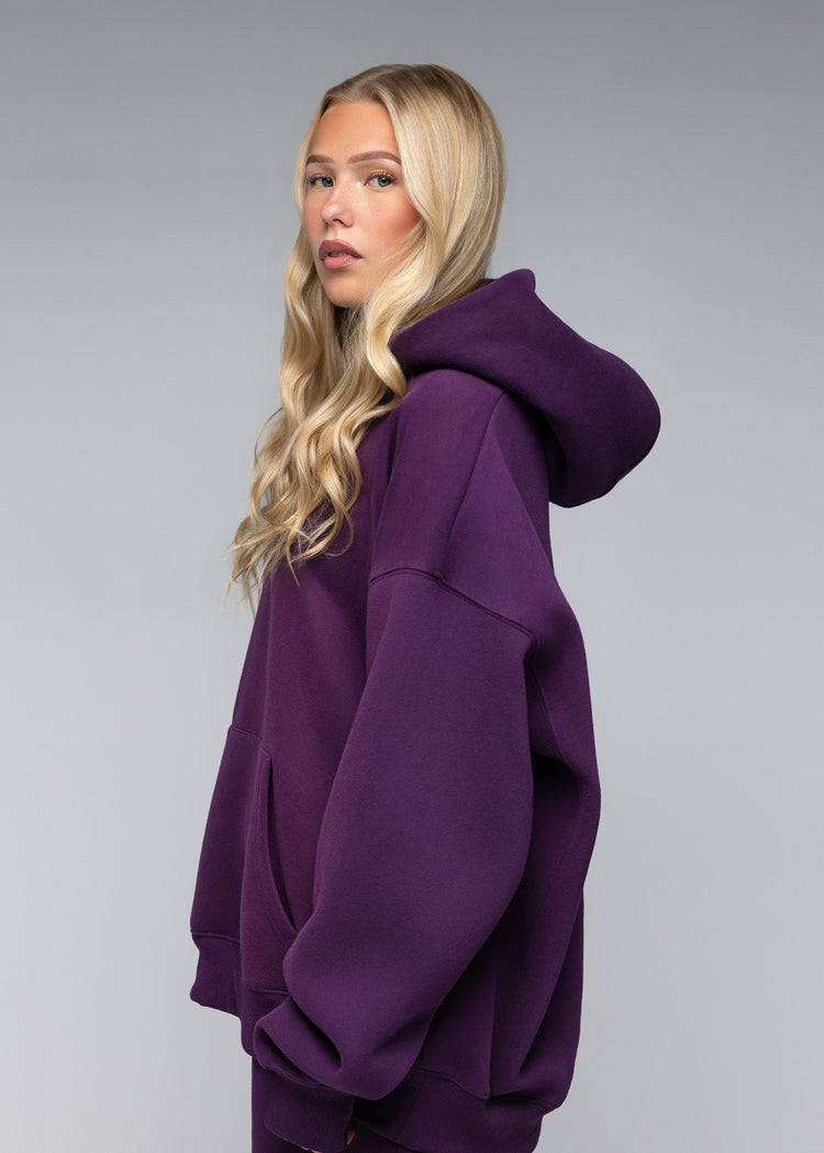 A blonde woman wearing an oversized deep purple hoodie and matching purple pants, photographed against a neutral gray studio background.