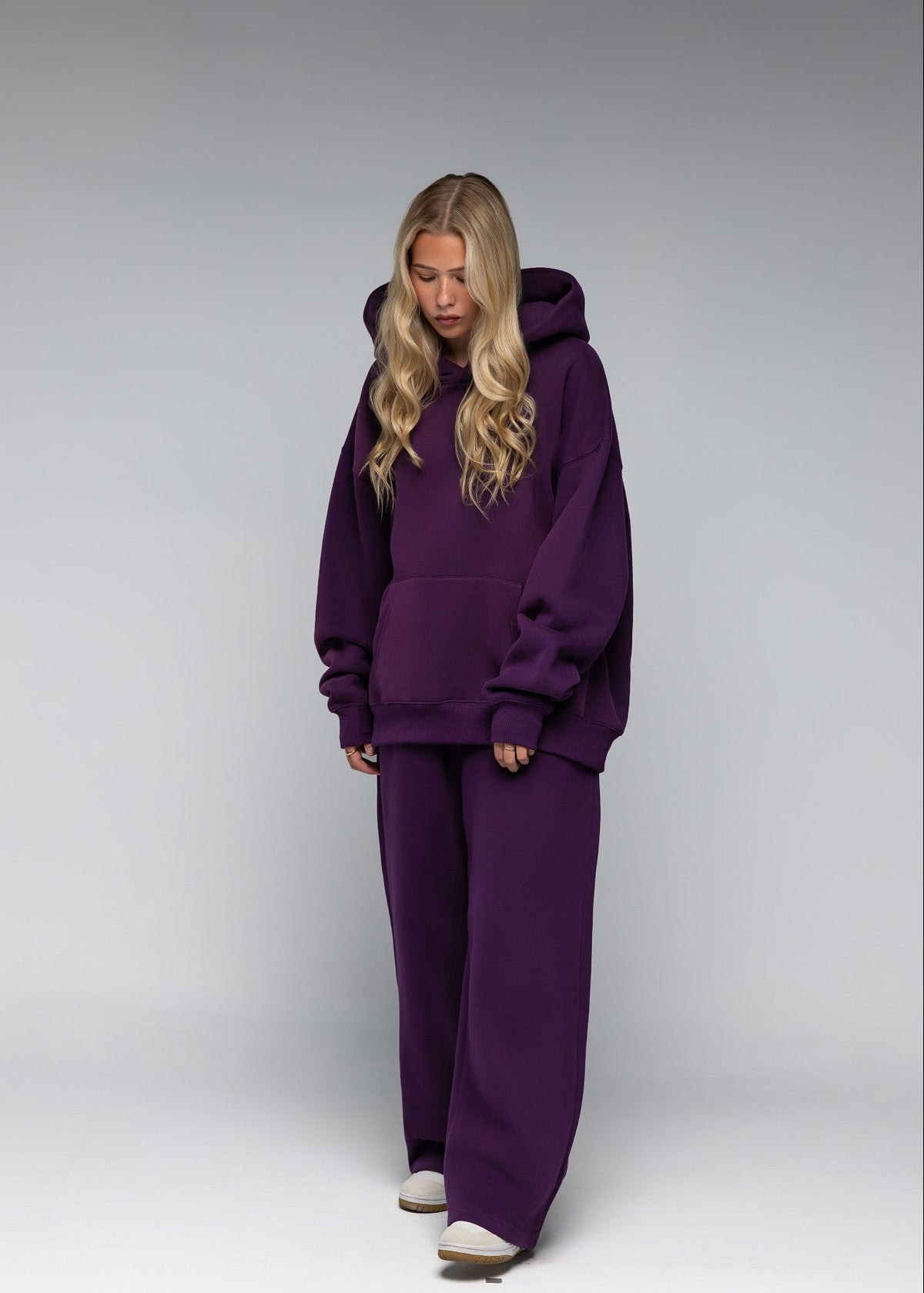 A woman modeling a matching deep purple lounge set consisting of a pullover hoodie and wide-leg sweatpants, standing against a neutral gray studio background.