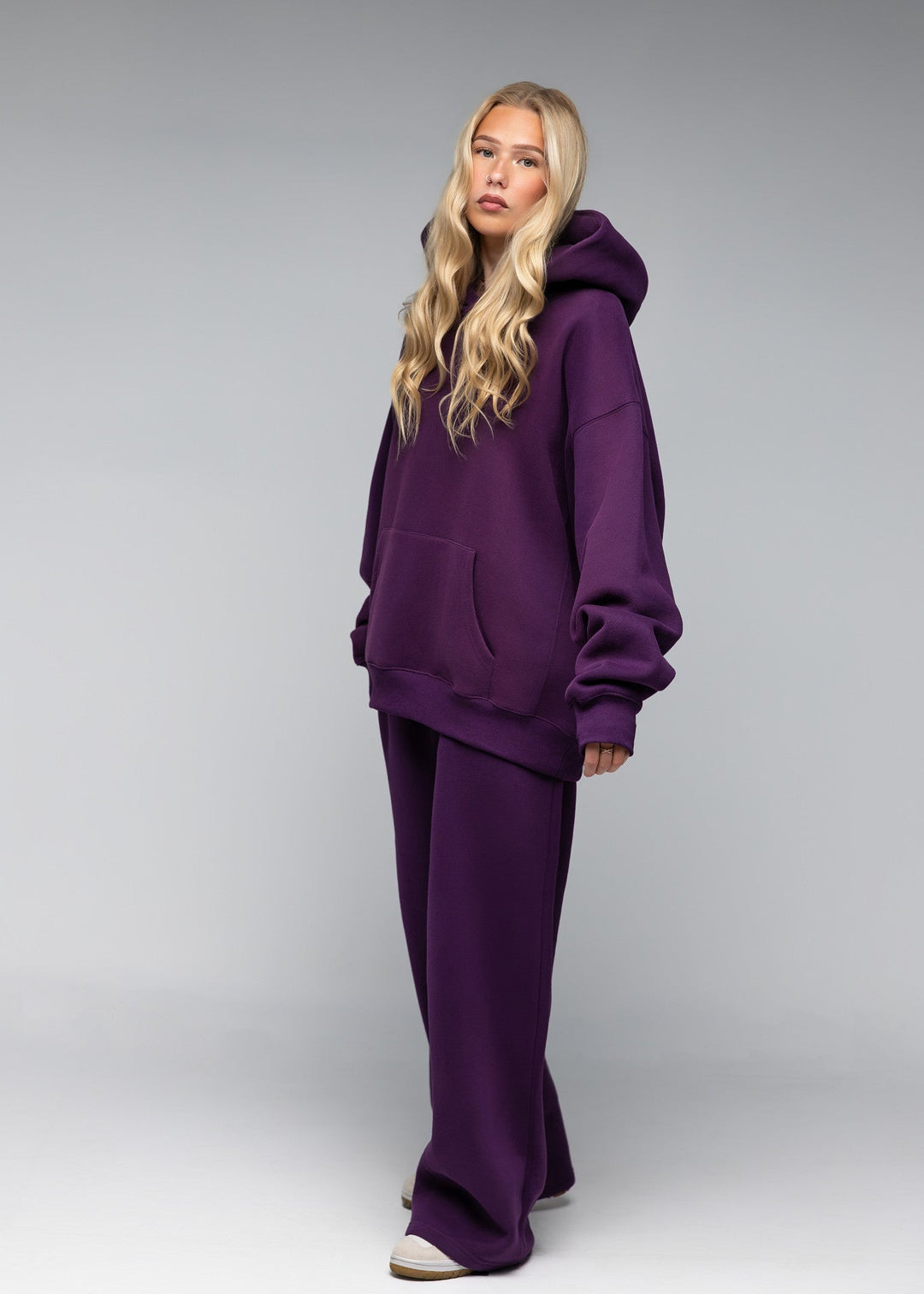 A blonde model wearing a matching deep purple lounge set consisting of an oversized pullover hoodie and wide-leg sweatpants, standing against a neutral gray studio background.