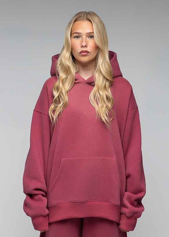 A blonde woman wearing an oversized dusty rose pink hoodie and matching sweatpants, standing against a neutral gray studio background.